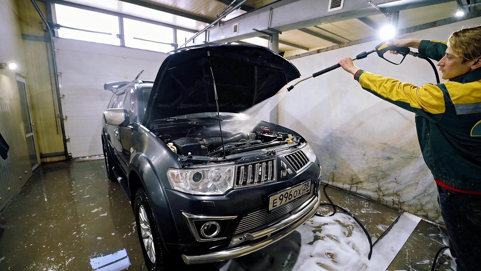 Мойщик моет моторный отсек внедорожника аппаратом высокого давления на посту автомойки